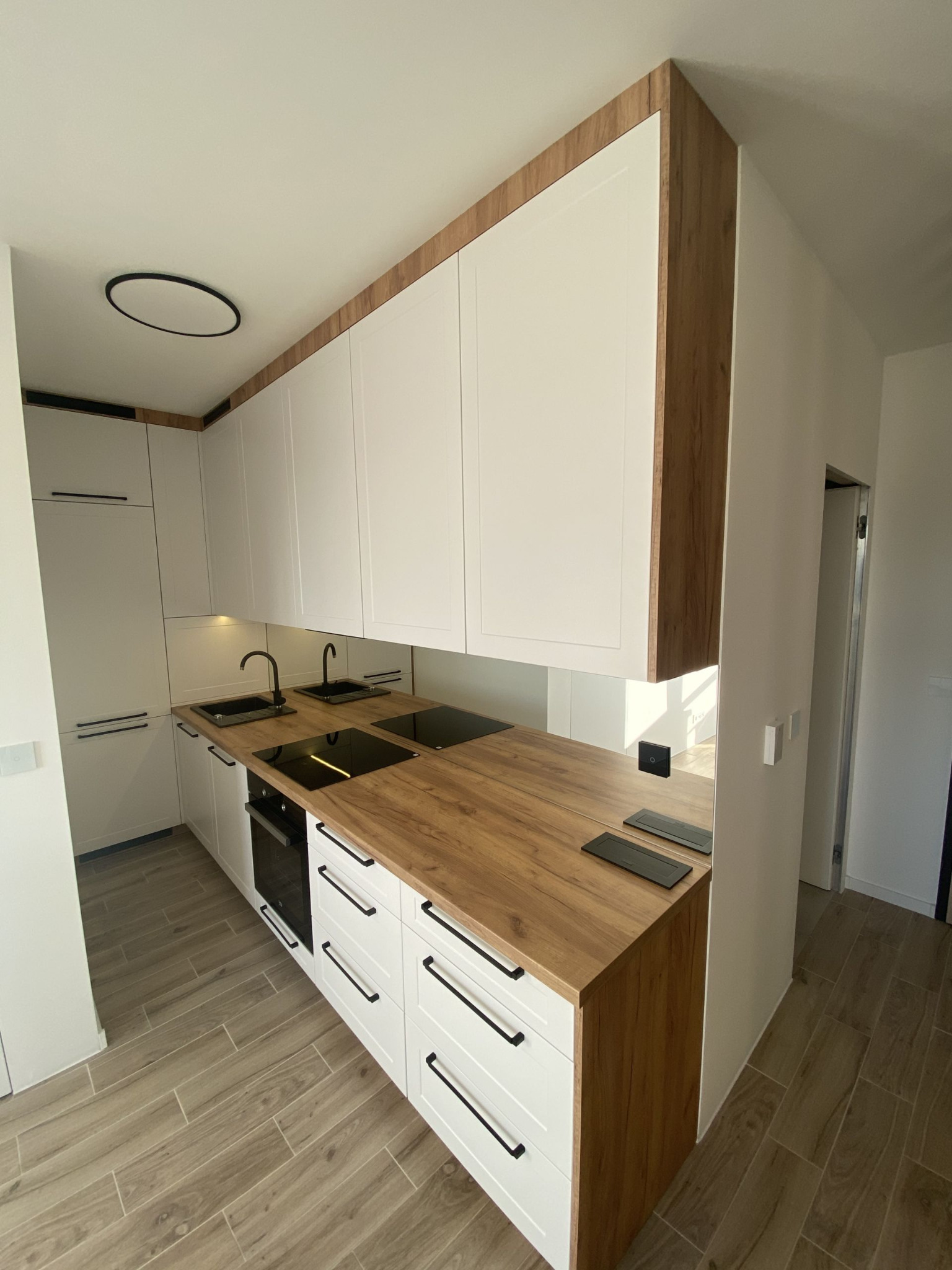 Nowoczesna kuchnia z białymi szafkami, drewnianym blatem i czarnymi akcentami, takimi jak bateria i płyta indukcyjna. Widoczne podświetlenie LED pod szafkami wiszącymi i drewniane wykończenie...