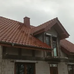 Dom w trakcie budowy z nowym, czerwonym dachem ceramicznym, widoczne elementy więźby i komin z drabinkami, szare niebo w tle.