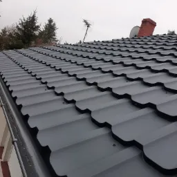 Nowy, grafitowy dach z blachodachówki modułowej, widoczny w zbliżeniu, z wkrętarką DeWalt na pierwszym planie, komin z czerwonej cegły w tle, pochmurne niebo.