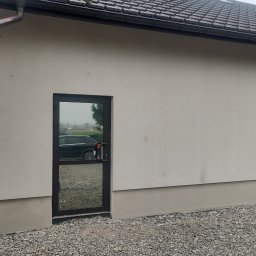 SolBud firma budowlana Krystian Solon - Elewacja budynku po tynkowaniu na jasny kolor, z widocznym cokołem w odcieniu szarości, okno z ciemną ramą odbija krajobraz i zaparkowany samochód, obok brązowe drzwi garażowe.