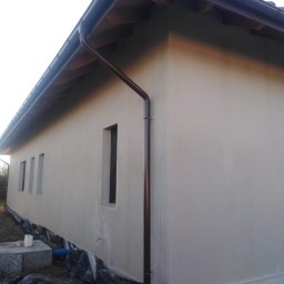 SolBud firma budowlana Krystian Solon - Świeżo otynkowana elewacja budynku jednorodzinnego w Mielcu, z brązowymi rynnami i trzema wąskimi oknami, widok z zewnątrz na tle przygotowanego terenu.