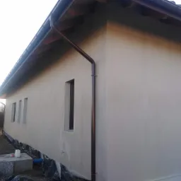 Świeżo otynkowana elewacja budynku jednorodzinnego w Mielcu, z brązowymi rynnami i trzema wąskimi oknami, widok z zewnątrz na tle przygotowanego terenu.