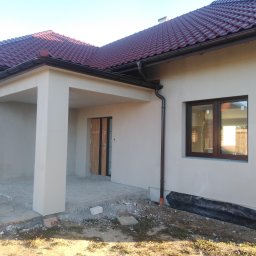 SolBud firma budowlana Krystian Solon - Elewacja domu jednorodzinnego po tynkowaniu, z widocznymi brązowymi oknami i częściowo ułożonym kamiennym chodnikiem przed wejściem.