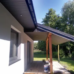 Nowoczesny dom jednorodzinny z wykończoną elewacją i zadaszonym tarasem, widoczne puszki z farbą na drewnianej podłodze tarasu, w tle zieleń drzew.