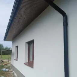 Elewacja domu po ociepleniu, widoczne brązowe okna, biały tynk, czarna rynna oraz podbitka dachowa w kolorze drewna.