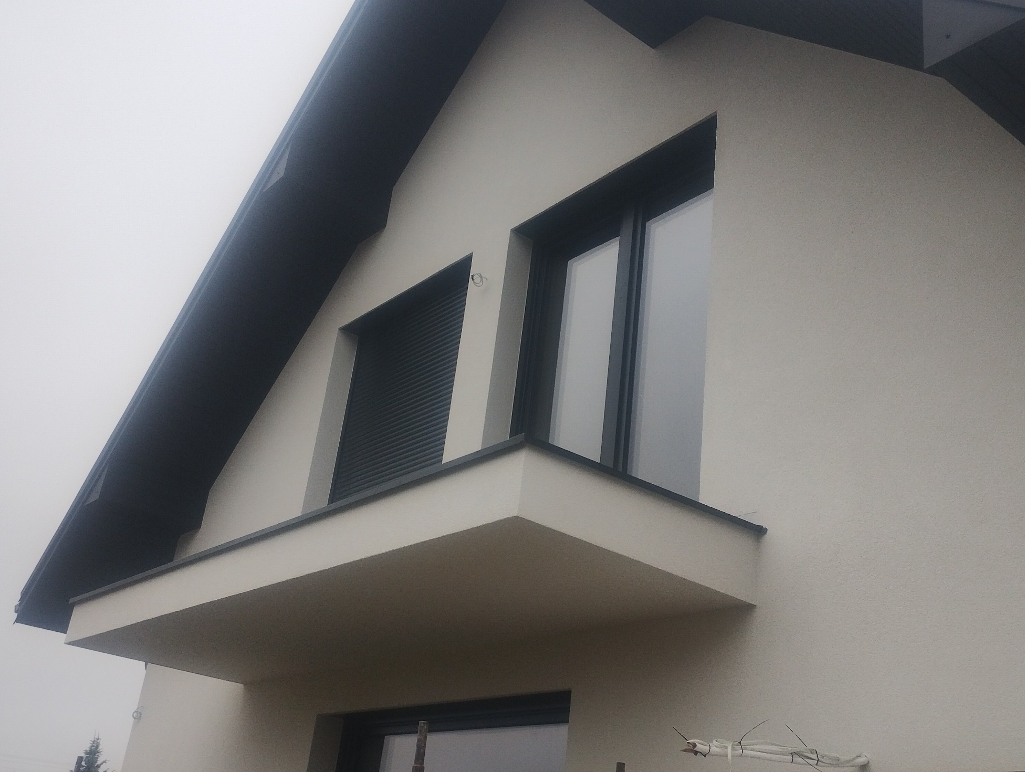 Nowoczesny dom z jasną elewacją, ciemnoszarą stolarką okienną i roletami zewnętrznymi, widok z dołu w pochmurny dzień.