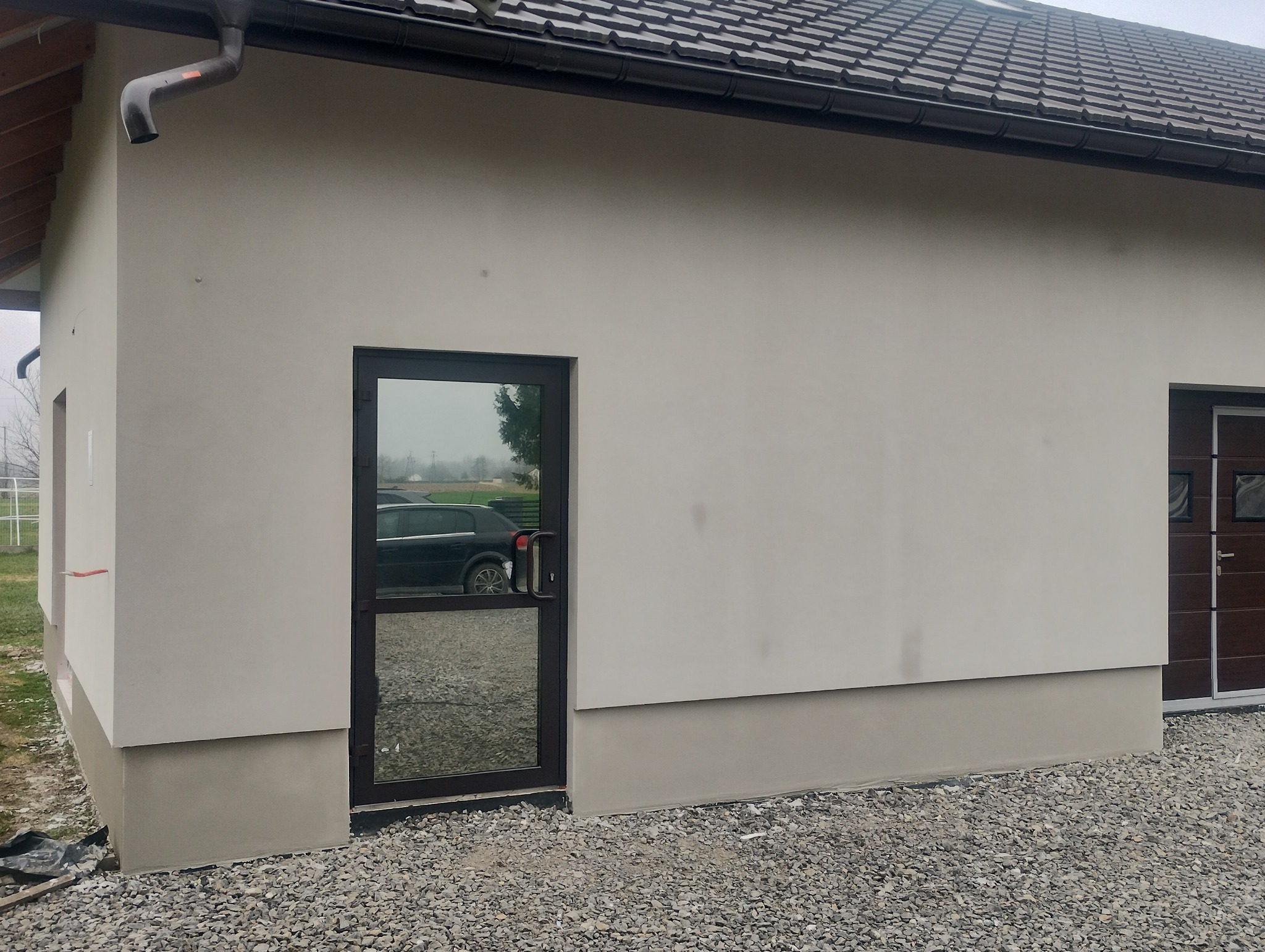 Elewacja budynku po tynkowaniu na jasny kolor, z widocznym cokołem w odcieniu szarości, okno z ciemną ramą odbija krajobraz i zaparkowany samochód, obok brązowe drzwi garażowe.