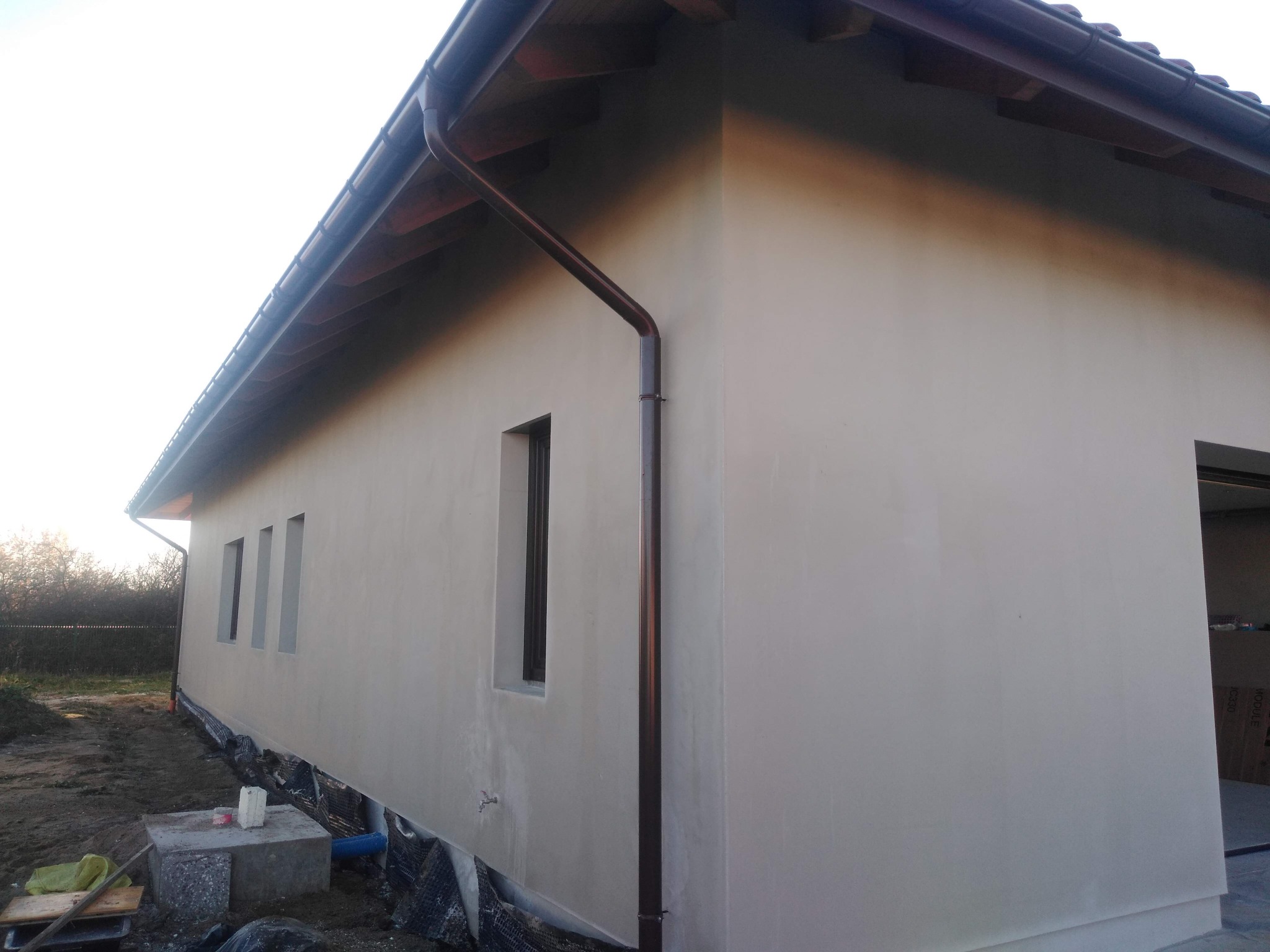 Świeżo otynkowana elewacja budynku jednorodzinnego w Mielcu, z brązowymi rynnami i trzema wąskimi oknami, widok z zewnątrz na tle przygotowanego terenu.