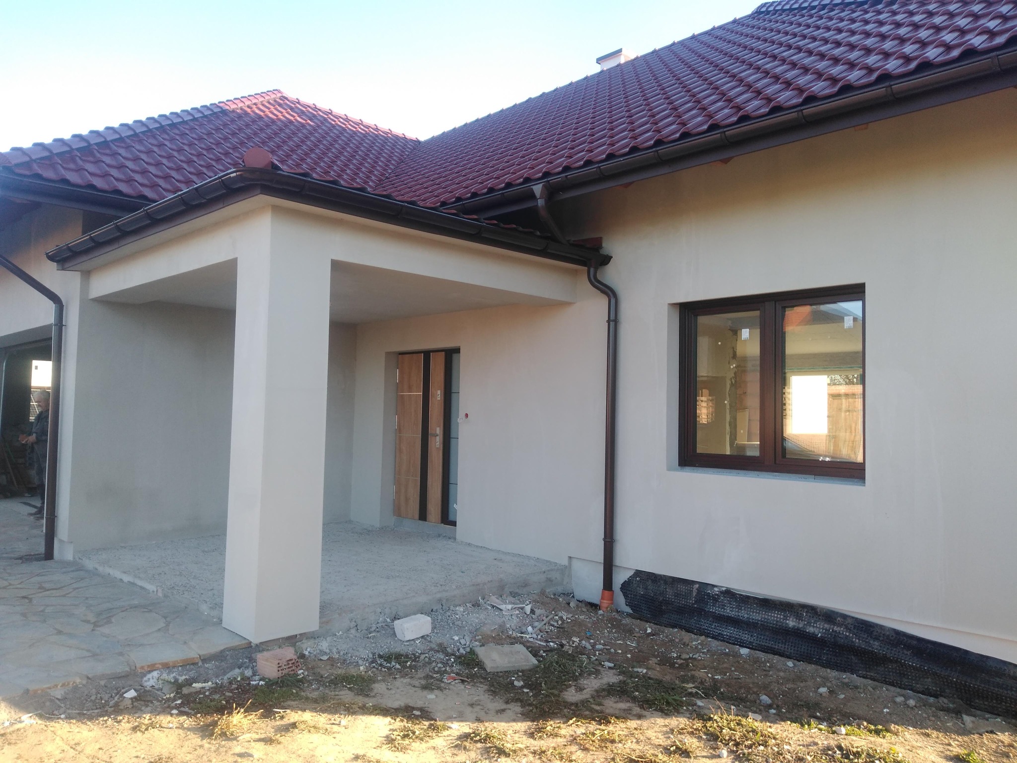 Elewacja domu jednorodzinnego po tynkowaniu, z widocznymi brązowymi oknami i częściowo ułożonym kamiennym chodnikiem przed wejściem.