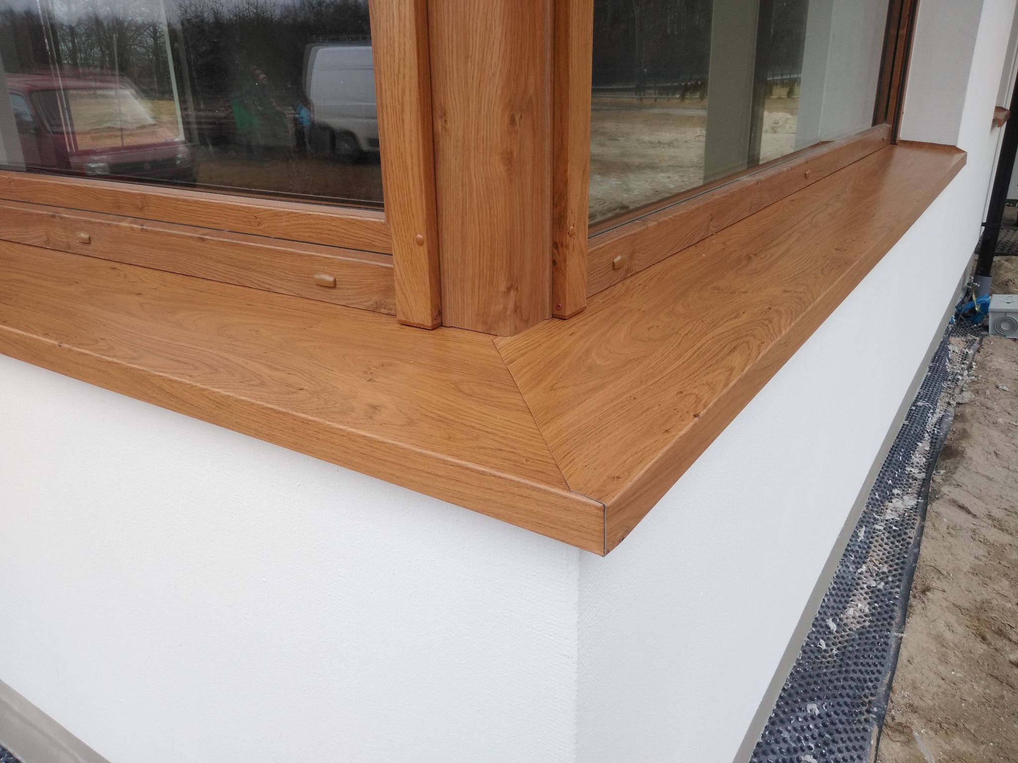 Fragment elewacji z zamontowanym oknem z drewnianą ramą i parapetem, widoczna warstwa ocieplenia przy gruncie.