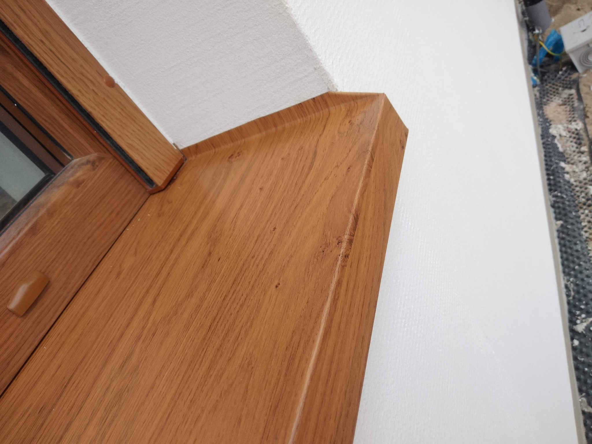 Detal wykończenia ościeżnicy okna z drewnopodobnym dekorem, połączonej z białą, świeżo pomalowaną ścianą. Widoczne ślady prac remontowych w tle.