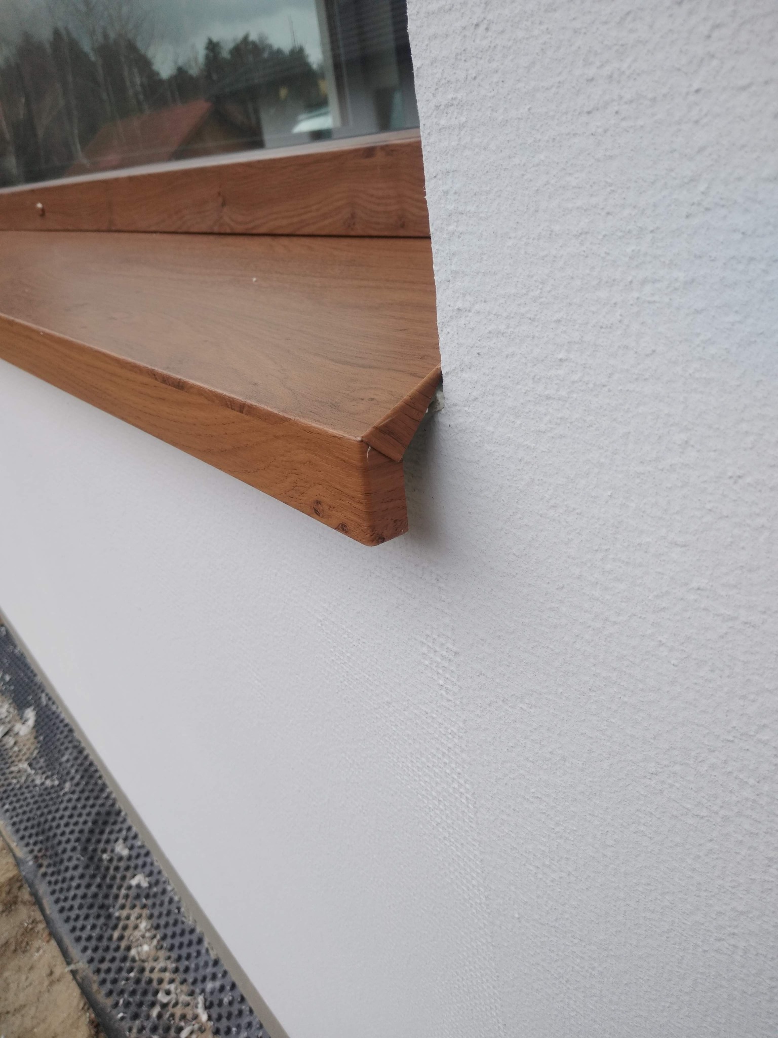 Nowy, drewnopodobny parapet zewnętrzny zamontowany na ocieplonej, białej elewacji budynku.