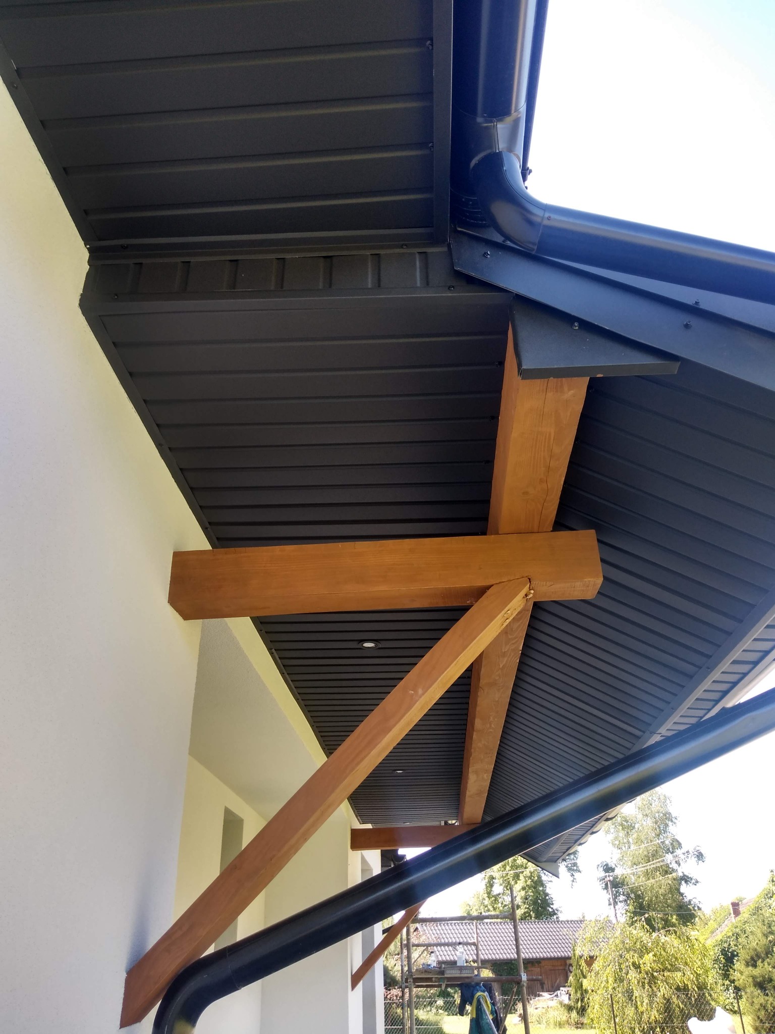 Zadaszenie wejścia do budynku z ciemnoszarej blachy trapezowej, wsparte na drewnianych belkach, z widoczną rynną w kolorze czarnym.