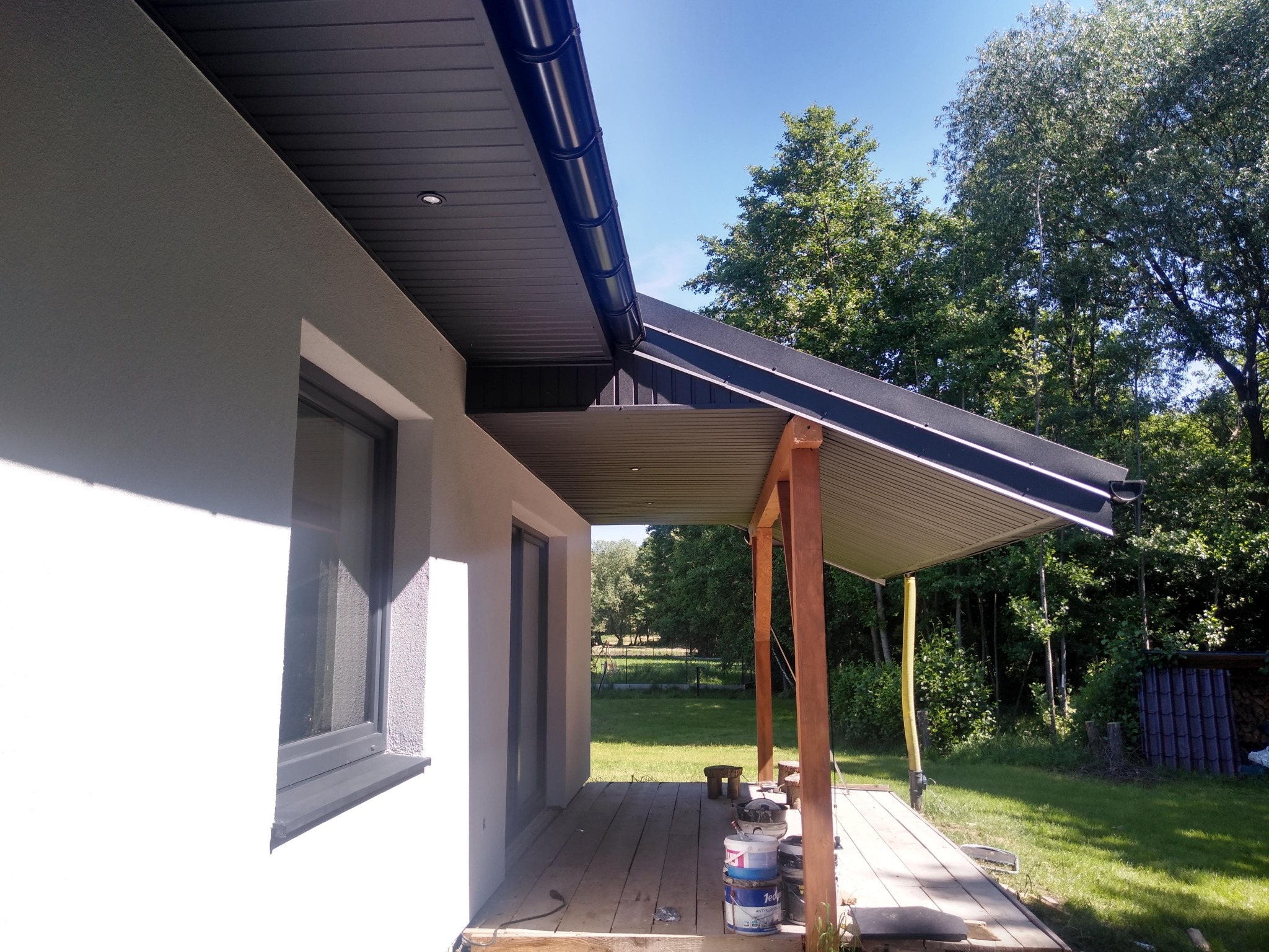 Nowoczesny dom jednorodzinny z wykończoną elewacją i zadaszonym tarasem, widoczne puszki z farbą na drewnianej podłodze tarasu, w tle zieleń drzew.