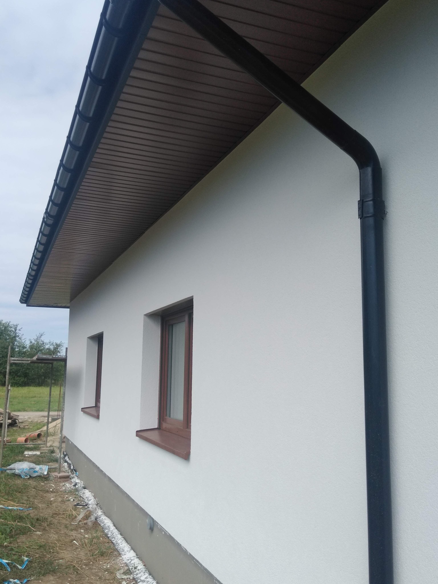 Elewacja domu po ociepleniu, widoczne brązowe okna, biały tynk, czarna rynna oraz podbitka dachowa w kolorze drewna.