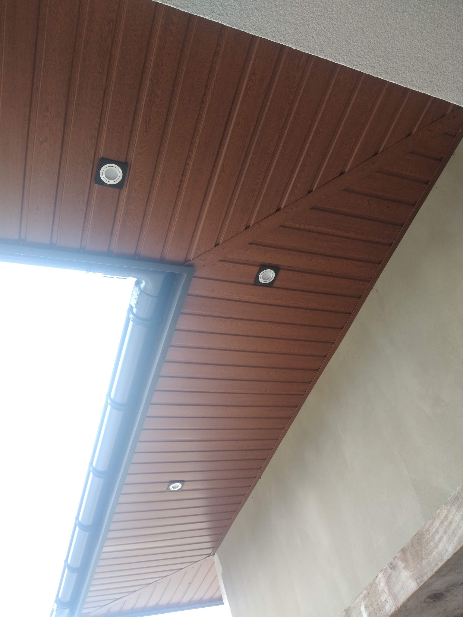 Podbitka dachowa z paneli PCV w kolorze brązowym, z wbudowanymi punktowymi lampami LED, widoczny fragment orynnowania i fragment ściany budynku.