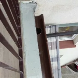 Szczegółowe ujęcie narożnika balkonu z brązową barierką i rynną, widoczne ślady malowania na ścianie i dziura w rynnie.