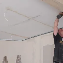 Pracownik na drabinie montuje drewnianą belkę do sufitu z płyt karton gips, używając wkrętarki akumulatorowej.