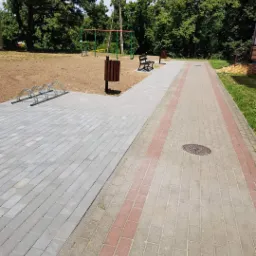 Nowo ułożona, szara kostka brukowa w parku, z widocznym stojakiem na rowery, koszem na śmieci i placem zabaw w tle.