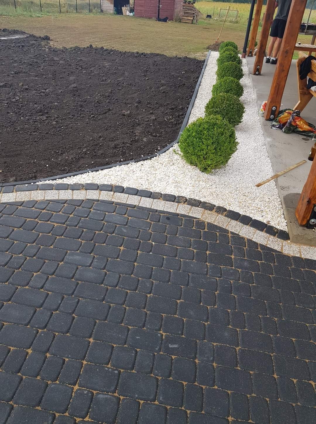 Ciemnoszara kostka brukowa ułożona łukiem, obrzeże z ciemnych kostek, biały żwir, kuliste krzewy i przygotowane podłoże pod trawnik na posesji.
