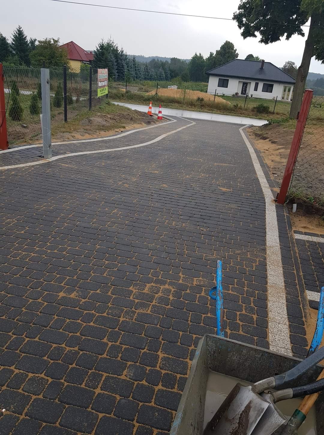 Czarna kostka brukowa ułożona na podjeździe z białymi liniami oddzielającymi, z widocznym w tle domem jednorodzinnym i narzędziami brukarskimi w pierwszym planie.