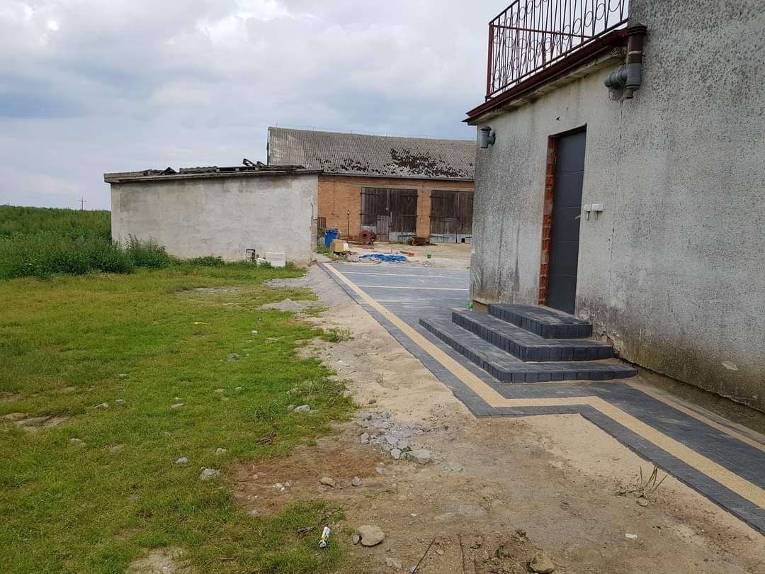 Ułożona kostka brukowa wzdłuż ściany budynku z schodami, widoczne trawnik i zabudowania gospodarcze w tle.