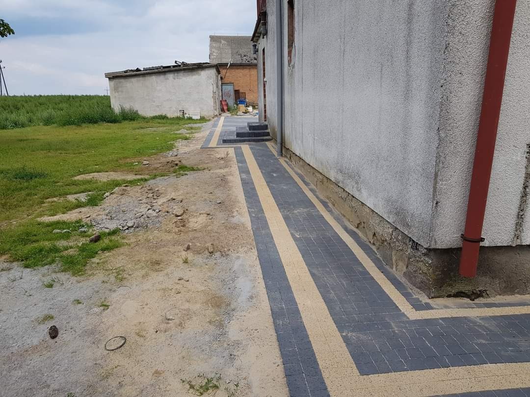 Ułożona ścieżka z kostki brukowej w kolorze szarym z beżowymi akcentami, biegnąca wzdłuż ściany budynku z czerwoną rurą spustową, z widocznym terenem przygotowywanym pod dalsze prace.