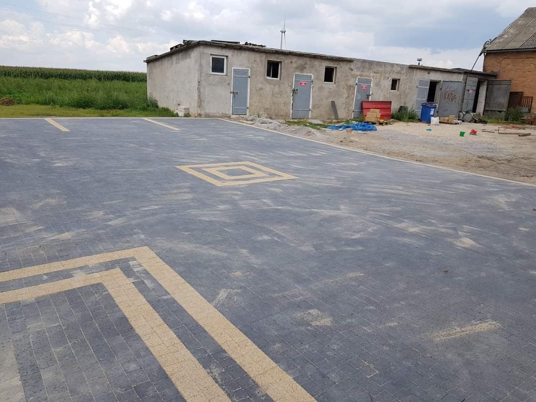 Nowo ułożona, szara kostka brukowa z geometrycznym wzorem w odcieniach beżu, otoczona budynkami gospodarczymi i polami w tle.