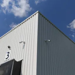 Biały magazyn z numerem 3 i zainstalowanymi kamerami monitoringu na tle błękitnego nieba z chmurami.