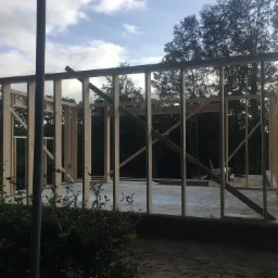 Wznoszenie drewnianej konstrukcji szkieletowej domu na betonowej płycie fundamentowej, widok przez krzewy, tło stanowią drzewa i pochmurne niebo.