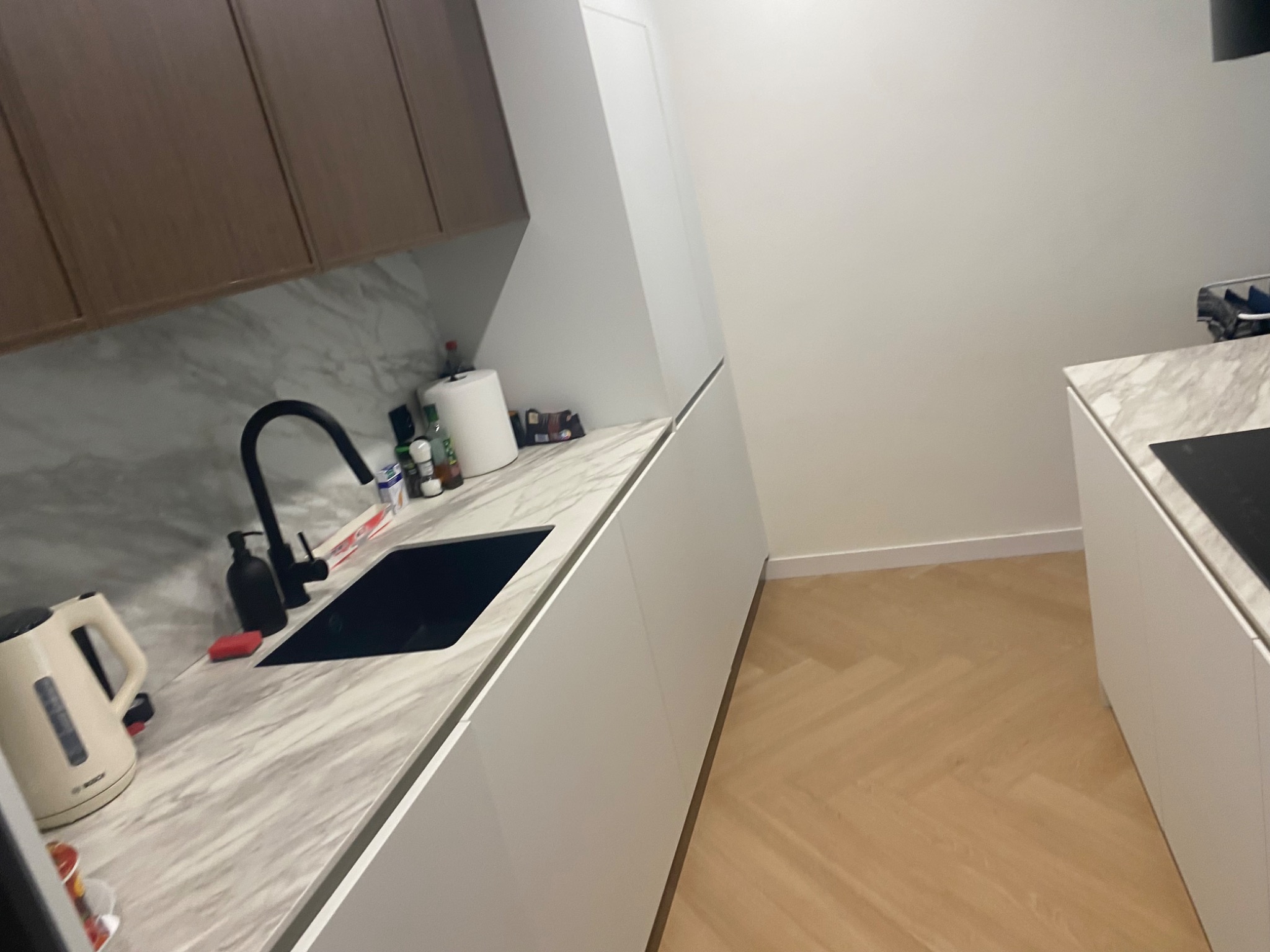 Nowoczesna kuchnia z marmurowym blatem, czarną armaturą i białymi szafkami. Parkiet w jodełkę. Minimalistyczny design, jasne wnętrze.