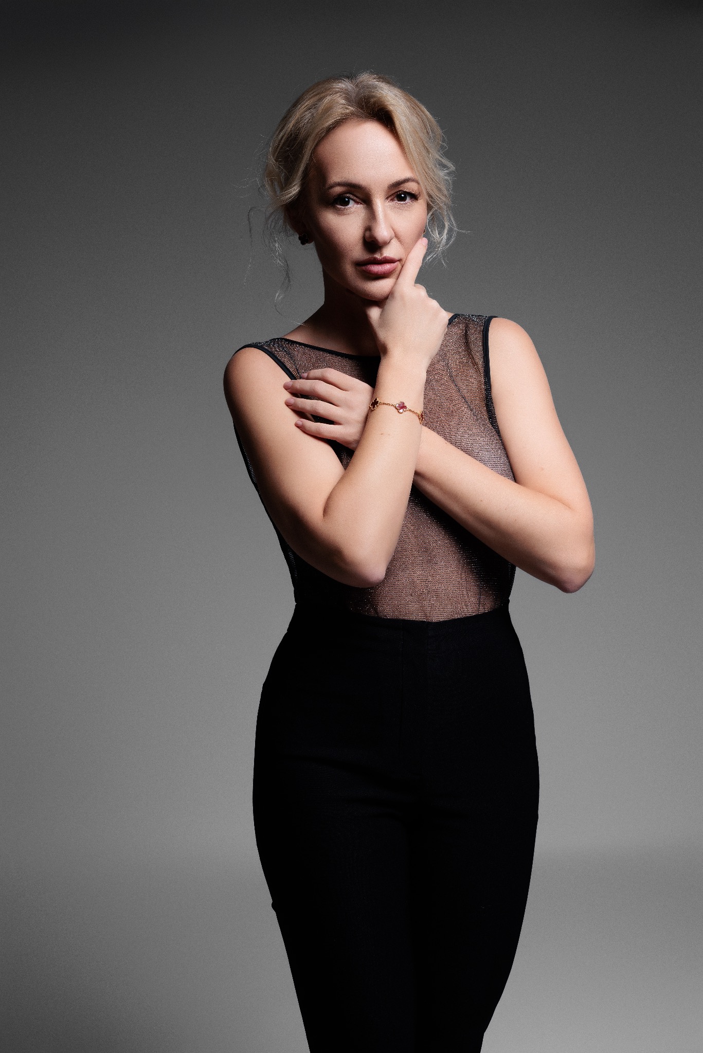 Portret kobiety w czarnej sukience na szarym tle, z ręką podpartą pod brodą i złotą bransoletką z czerwonymi kamieniami.