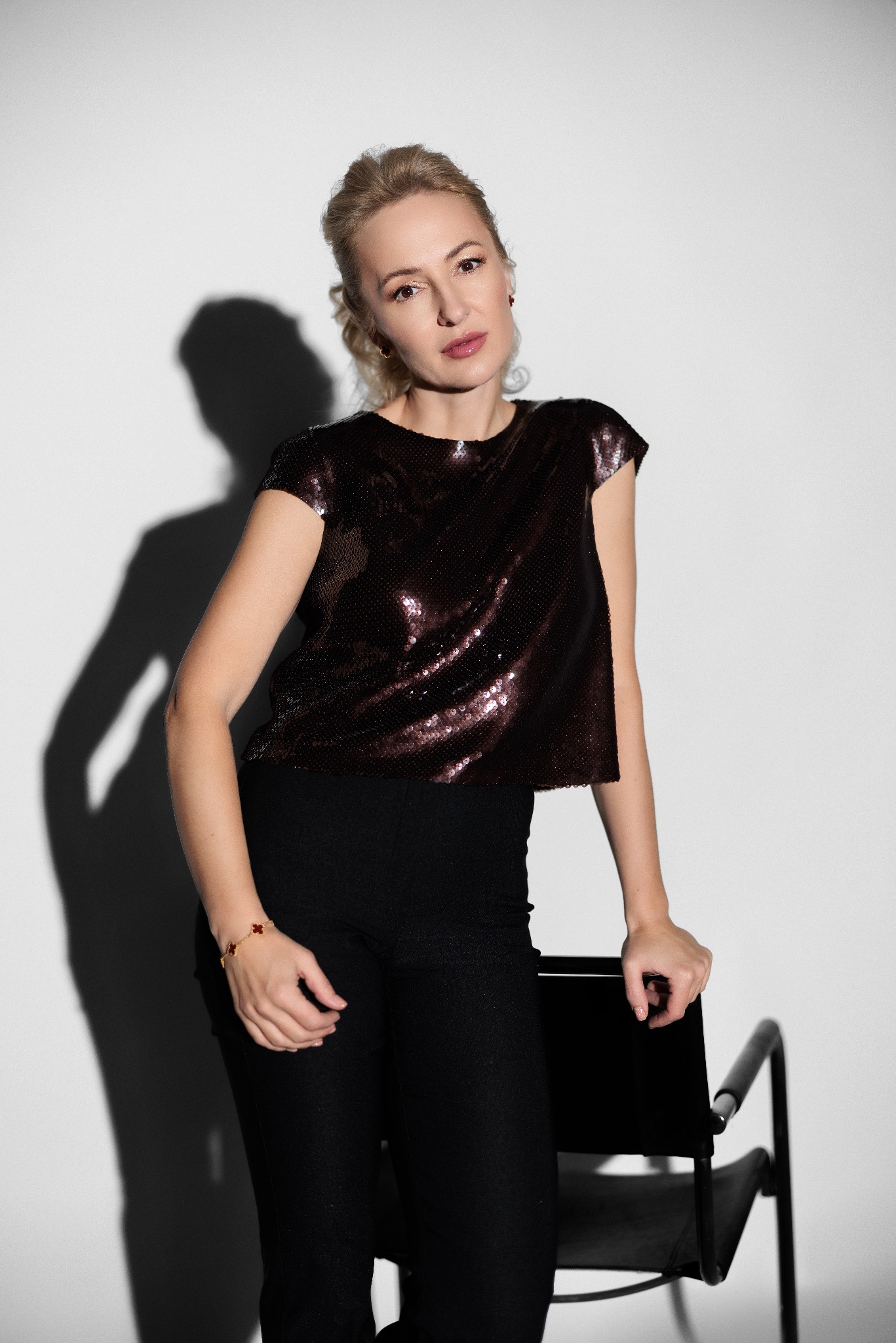 Portret kobiety w cekinowej bluzce i czarnych spodniach, opierającej się o czarne krzesło na jasnym tle, z widocznym cieniem.