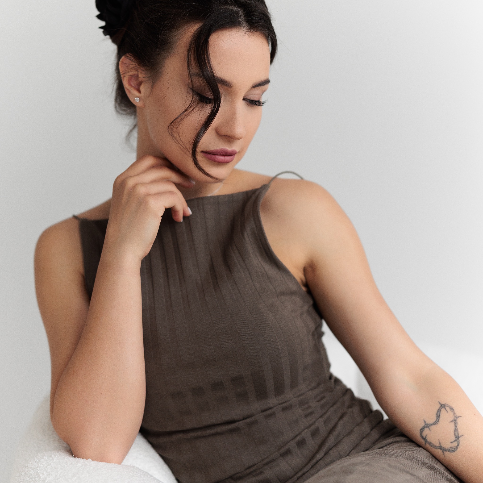 Portret kobiety w prążkowanej sukience, z delikatnym tatuażem serca z drutu kolczastego na ramieniu, siedzącej na białym fotelu i spoglądającej w dół.