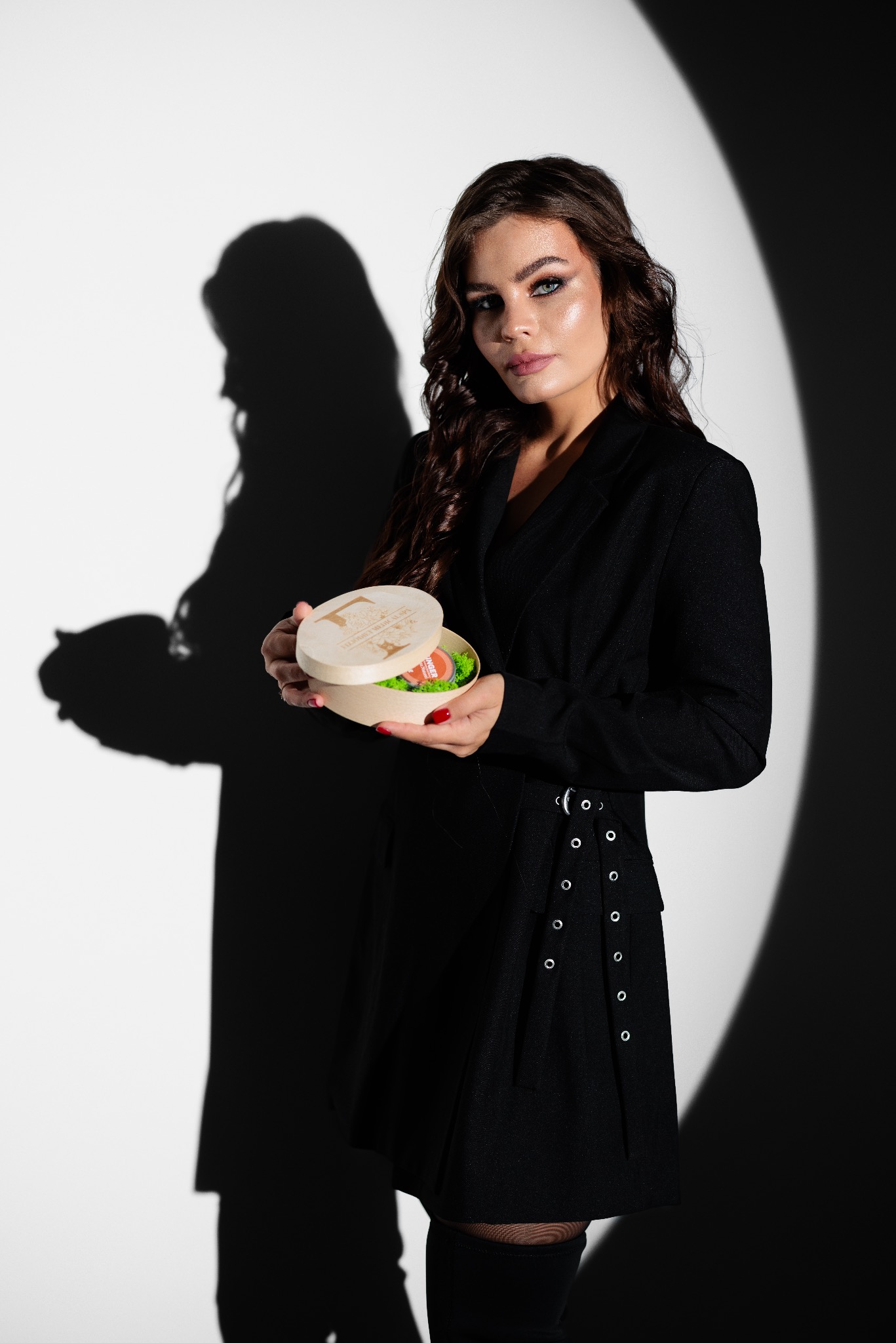 Kobieta w czarnym żakiecie trzyma otwarte, okrągłe pudełko z drewnianym wieczkiem, wypełnione zielonym wypełniaczem i produktem, na tle białej ściany z cieniem sylwetki.