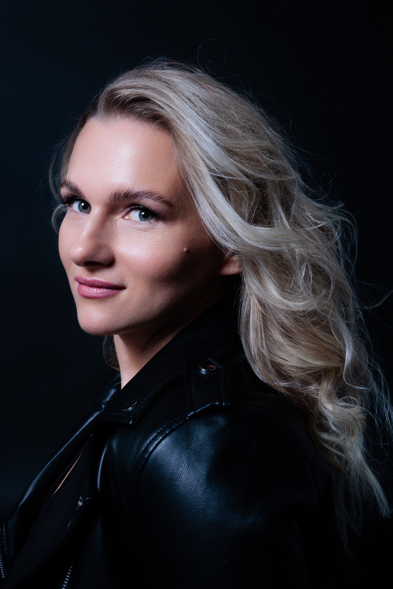 Portret kobiety z blond włosami w skórzanej kurtce na ciemnym tle, uśmiechająca się, patrzy w lewo.