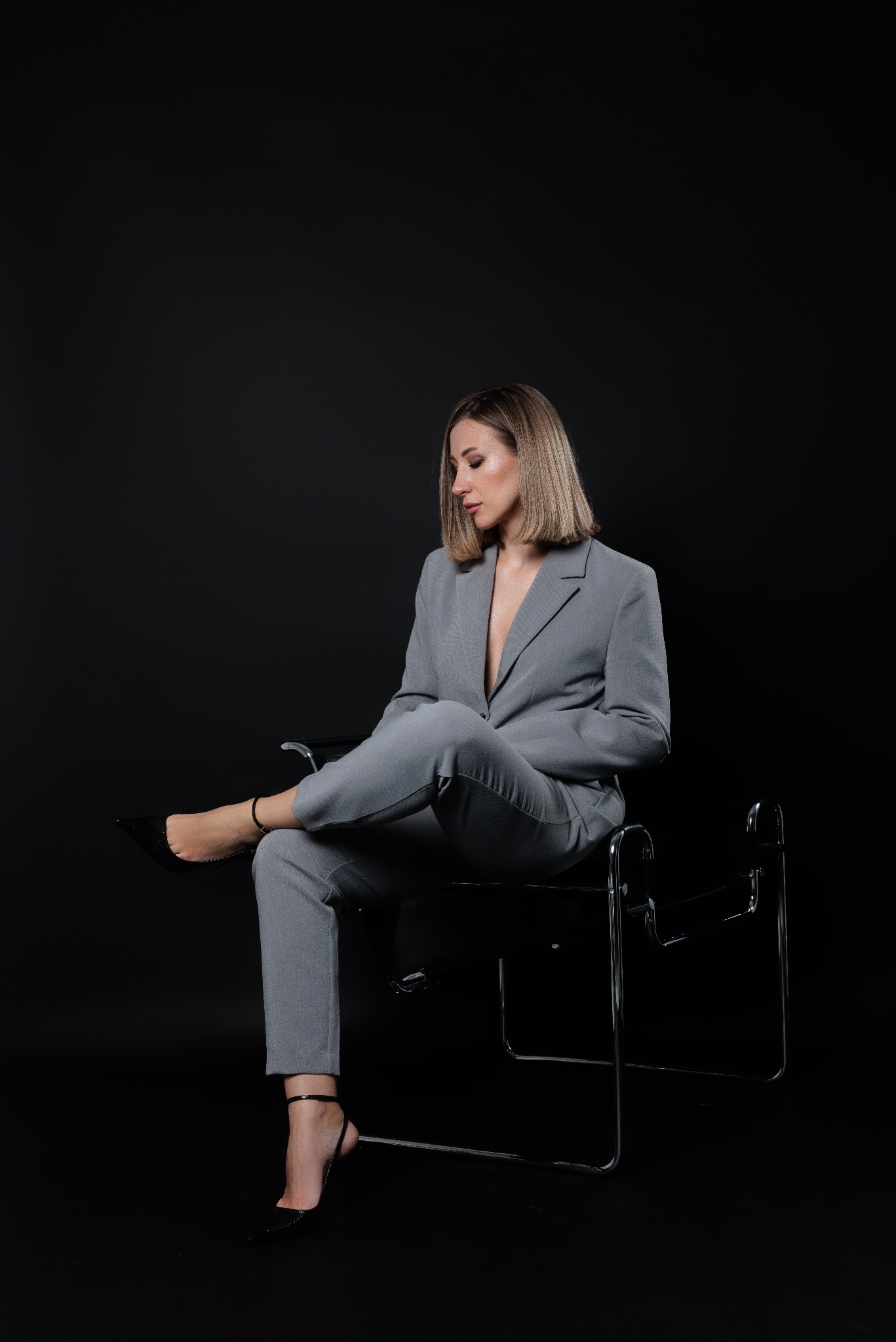 Kobieta w szarym garniturze siedzi na czarnym krześle na czarnym tle, patrząc w dół