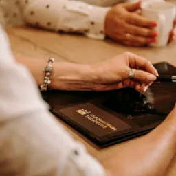 Kobieta trzyma długopis nad czarnym folderem z napisem 'Laboratorium Podatkowe', druga osoba w tle pije kawę.