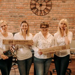 Laboratorium Podatkowe Tax-Lab Tarnów - Cztery uśmiechnięte kobiety w biurze z ceglaną ścianą i dużym zegarem, trzymające drewniane tabliczki z nazwami firm 'Laboratorium Podatkowe' i 'Tax-Lab'. Jedna z nich trzyma kalkulator.