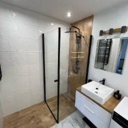 Nowoczesna łazienka z kabiną prysznicową typu walk-in z czarnymi ramami, białą umywalką nablatową na drewnianym blacie i białymi płytkami ściennymi o subtelnej fakturze.