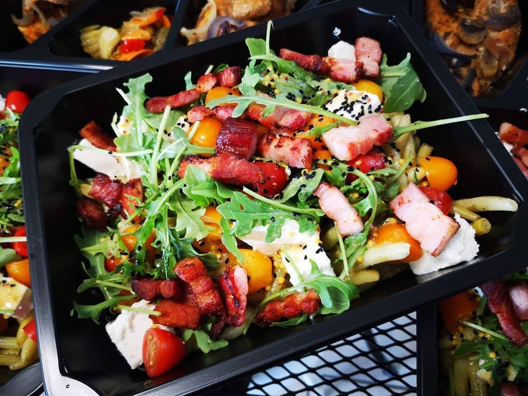 Sałatka z rukolą, pomidorkami koktajlowymi, serem tofu i chrupiącym boczkiem w czarnym opakowaniu cateringowym, posypana czarnym sezamem i sosem musztardowym.