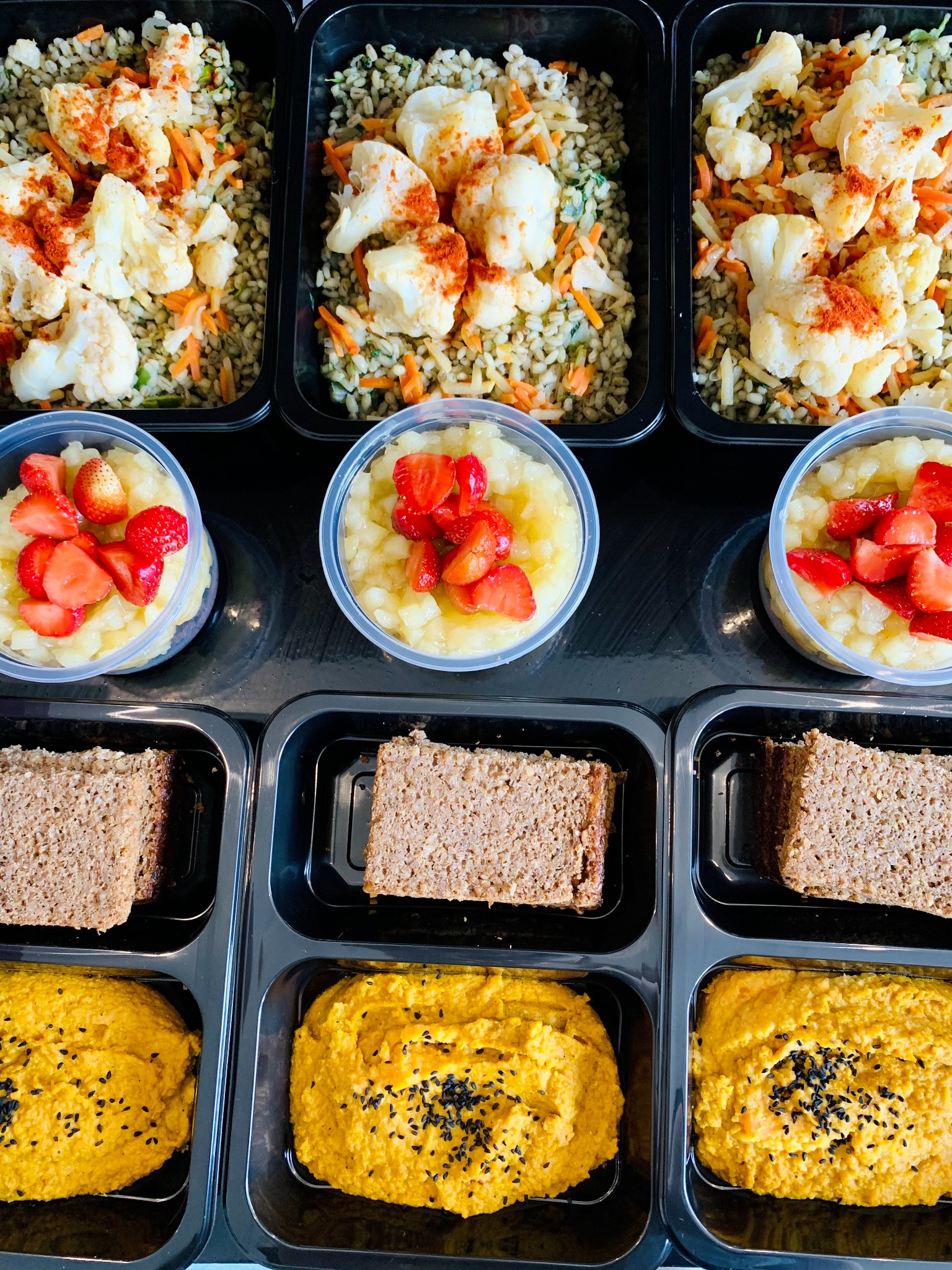 Zestaw posiłków w czarnych pojemnikach: kasza z warzywami i kalafiorem posypanym papryką, mus jabłkowy ze świeżymi truskawkami, kromka chleba pełnoziarnistego i hummus z czarnym sezamem.