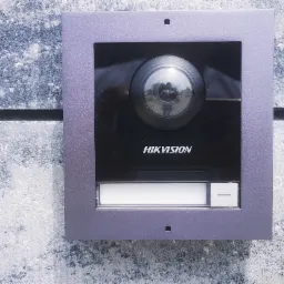 Zewnętrzny panel domofonu z kamerą Hikvision zamontowany na szarej, teksturowanej ścianie budynku. Widoczne odbicie otoczenia w obiektywie kamery.