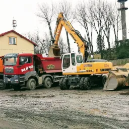 Żółto-biała koparka Liebherr 904 załadowuje urobek do czerwonej wywrotki MAN na placu budowy, w tle budynek i wieża.