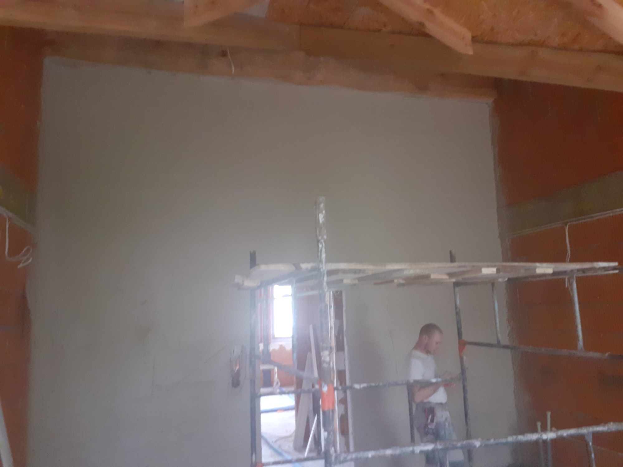 Świeżo otynkowana ściana w surowym wnętrzu poddasza, widoczny pracownik na rusztowaniu i elementy drewnianej konstrukcji dachu.