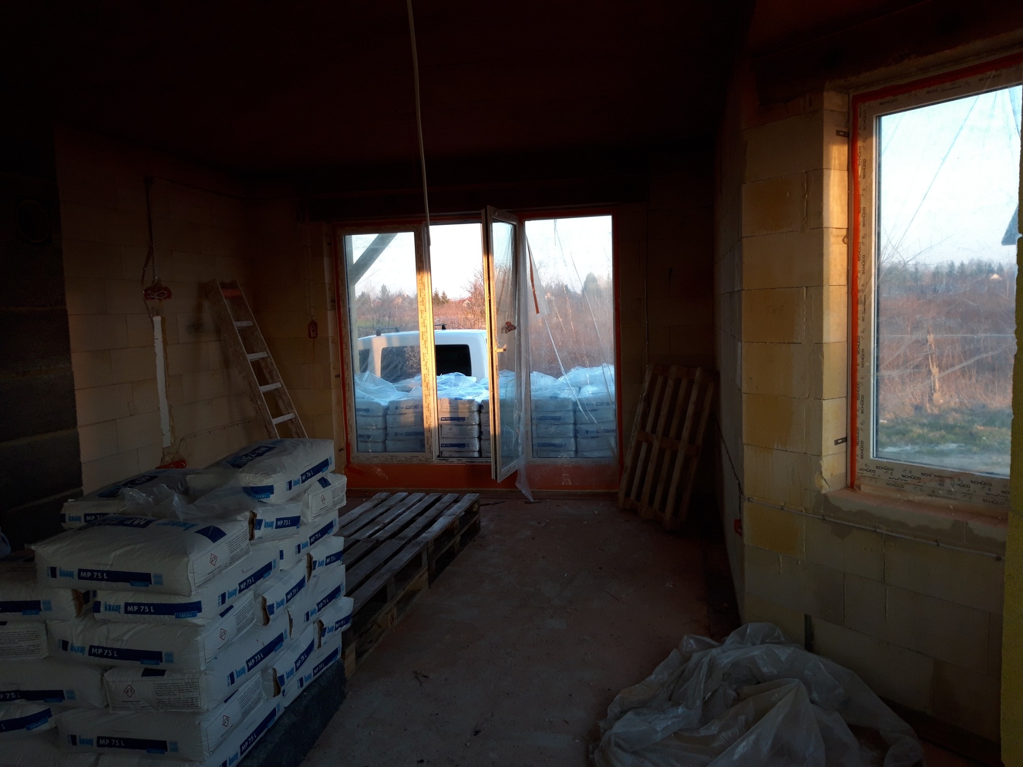 Wnętrze w stanie surowym przed tynkowaniem maszynowym: widoczne worki z zaprawą tynkarską Knauf MP 75 L na paletach, surowe ściany z pustaków, okna z widokiem na zewnątrz i drabina oparta o ścianę.