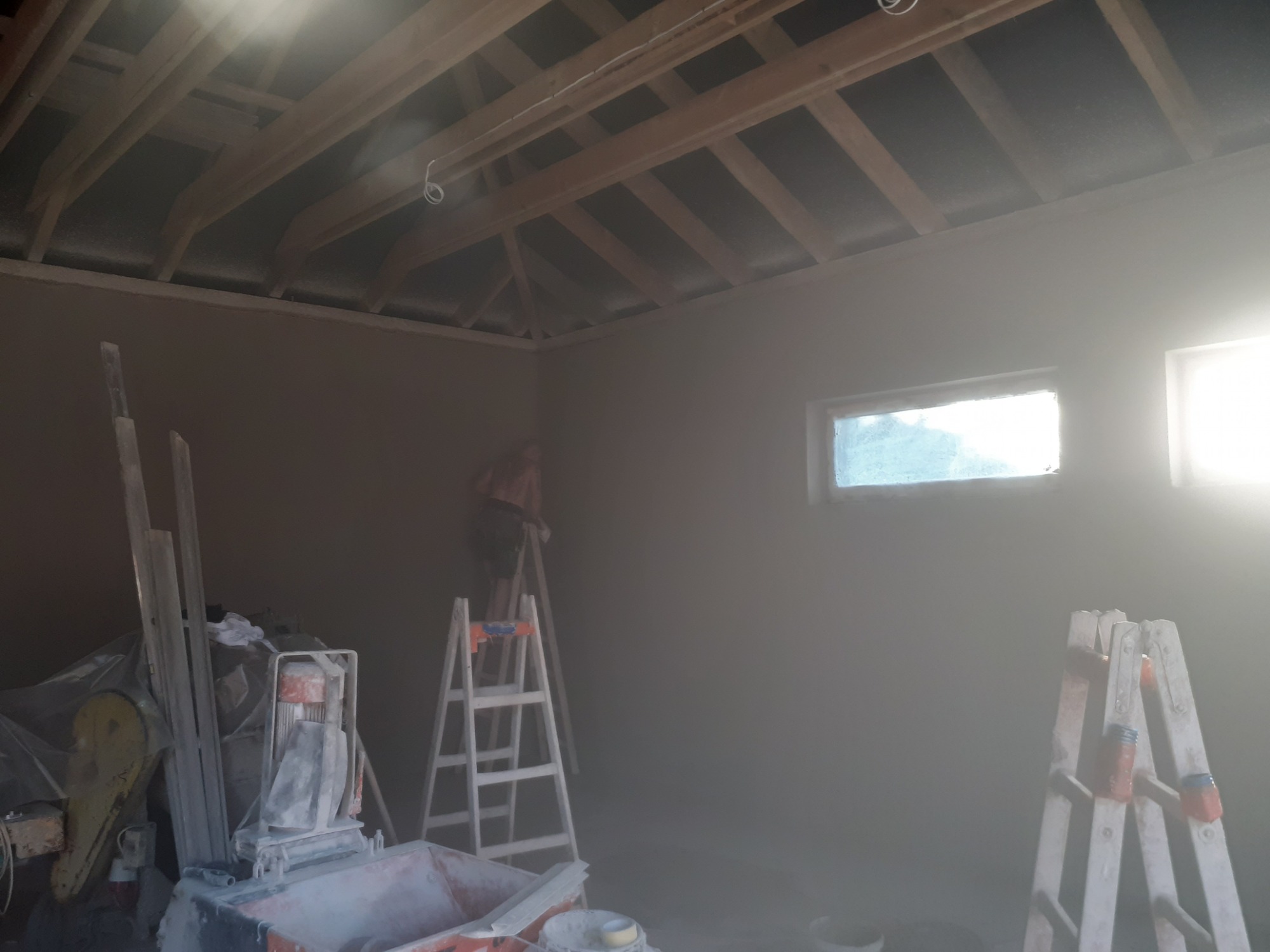 Wnętrze w trakcie nakładania tynku maszynowego, widoczna maszyna do tynkowania, drabina i pracownik na drabinie, surowe drewniane belki stropowe.