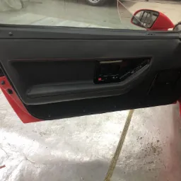 Czerwone auto z otwartymi drzwiami, ukazując czarną, skórzaną tapicerkę drzwi z czerwonymi przeszyciami, widoczne elementy sterowania szybą i zamkiem.