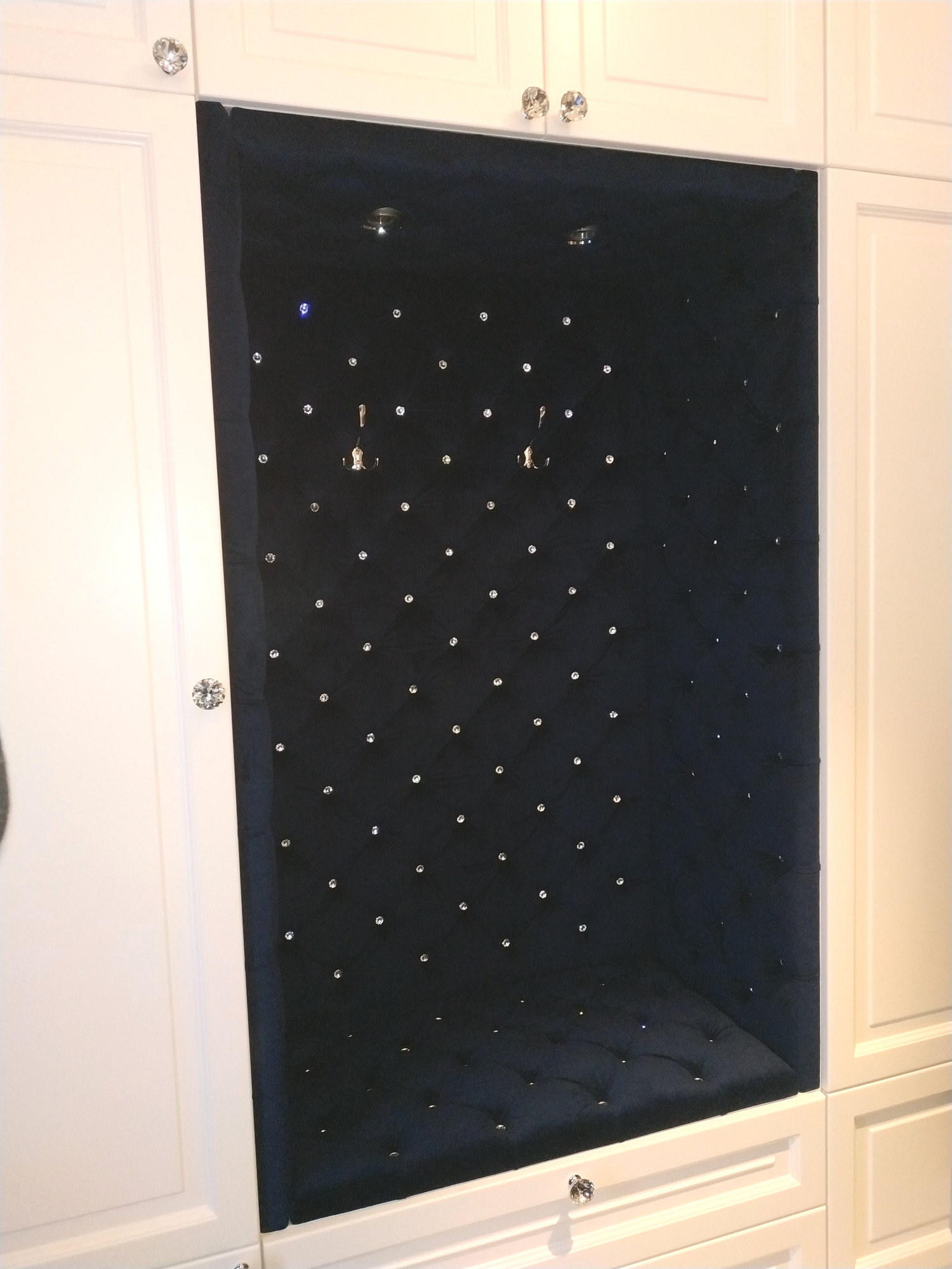 Wnęka w zabudowie meblowej z ciemnym, pikowanym obiciem z dekoracyjnymi kryształkami, oświetlona punktowo.