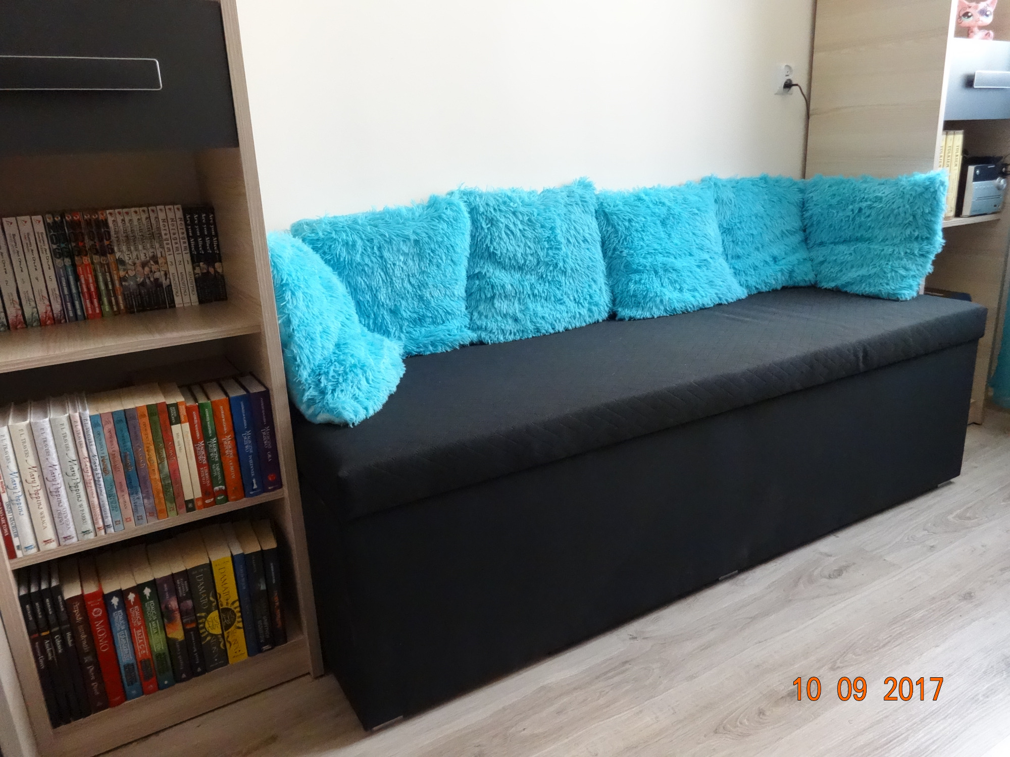 Tapicerowana ławka z czarnym siedziskiem i sześcioma turkusowymi poduszkami z długim włosiem, stojąca w pokoju obok regału z książkami.
